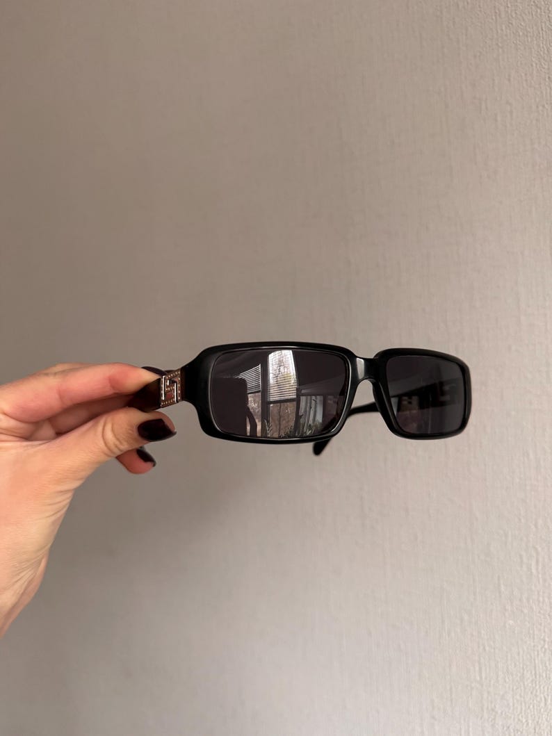 May include: Black rectangular sunglasses with dark lenses. The sunglasses have a thick frame and a silver accent on the temples. The lenses reflect the surrounding environment. The sunglasses are held by a hand.