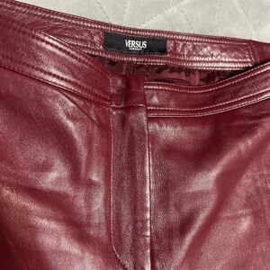 May include: Close-up of burgundy leather pants. The pants have a visible seam down the front and a black tag at the waistband with the text "VERSUS VERSACE". The leather has a smooth, shiny appearance.