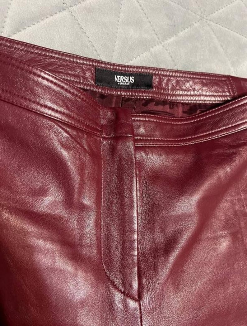 May include: Close-up of burgundy leather pants. The pants have a visible seam down the front and a black tag at the waistband with the text "VERSUS VERSACE". The leather has a smooth, shiny appearance.