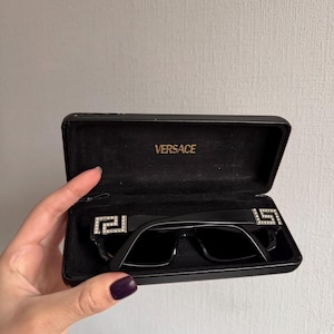 May include: Black sunglasses with a rectangular frame and dark lenses, displayed in an open black case. The sunglasses feature a decorative silver emblem on each temple. The case is embossed with the brand name "VERSACE".