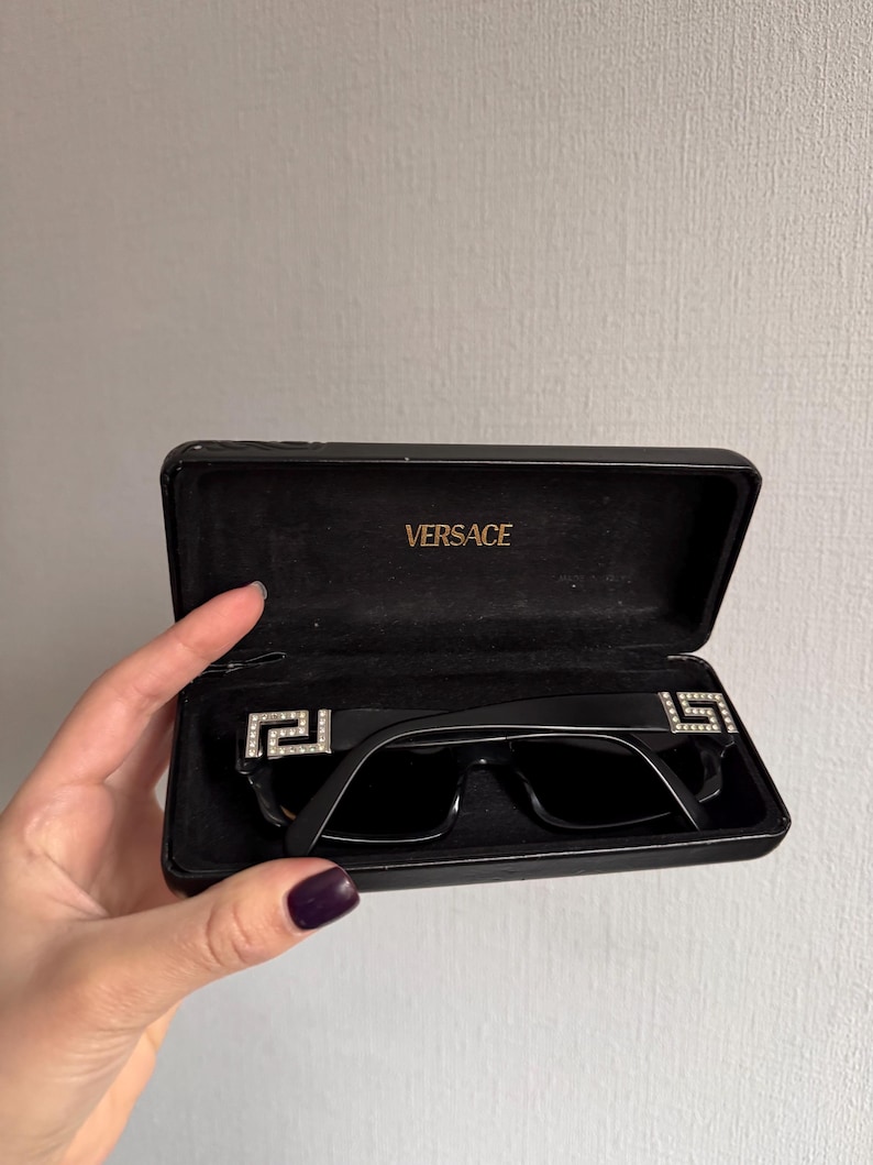 May include: Black sunglasses with a rectangular frame and dark lenses, displayed in an open black case. The sunglasses feature a decorative silver emblem on each temple. The case is embossed with the brand name "VERSACE".
