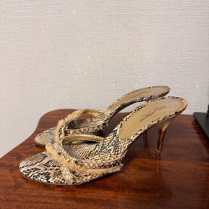 May include: Dolce & Gabbana high-heeled sandals with a snakeskin pattern in brown, black, and beige. The sandals have thin straps with fringe and slender, gold-toned heels. The shoes are on a wooden surface.