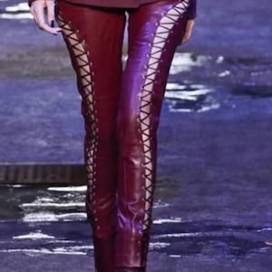 May include: Burgundy leather pants with lace-up detailing on the sides. The pants are paired with matching burgundy boots. The model is wearing a burgundy blazer. The outfit is on a runway.