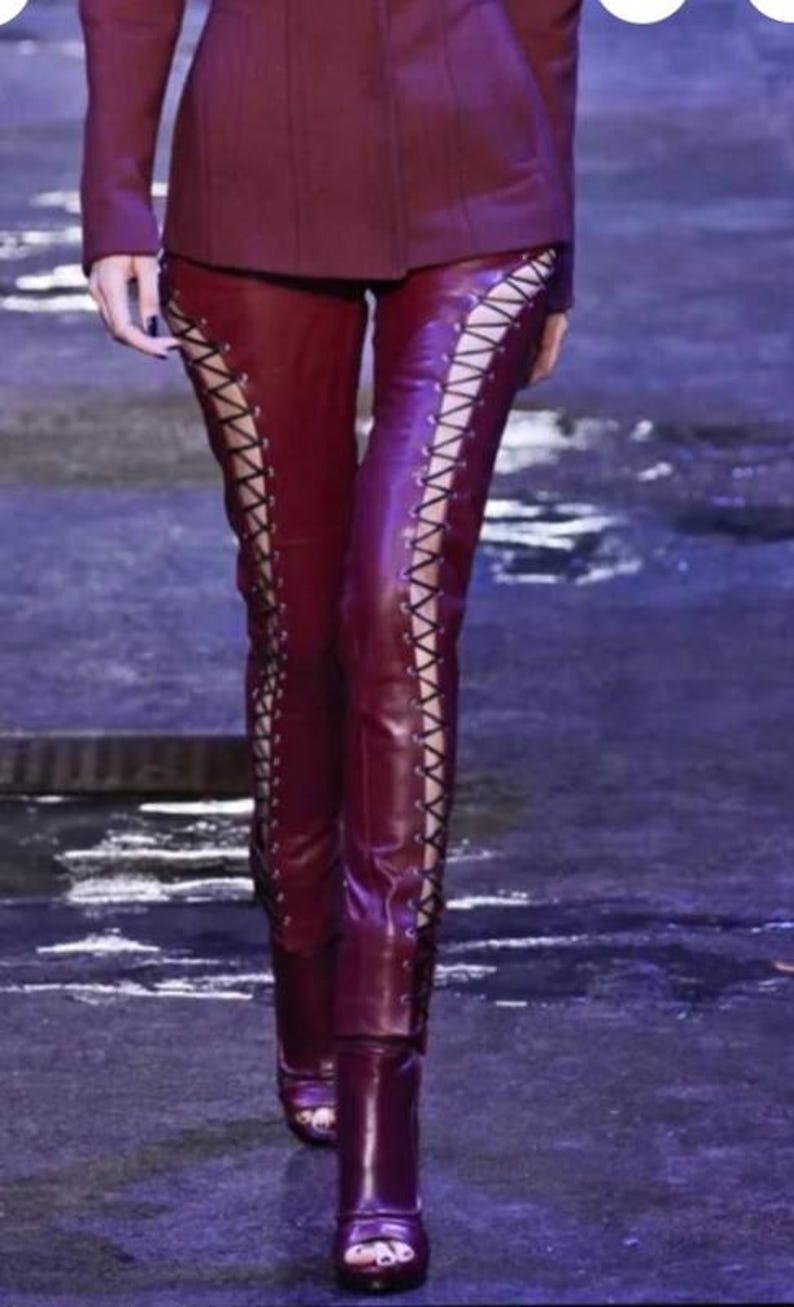 May include: Burgundy leather pants with lace-up detailing on the sides. The pants are paired with matching burgundy boots. The model is wearing a burgundy blazer. The outfit is on a runway.