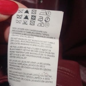 May include: A close-up of a burgundy garment tag with care symbols and text in multiple languages. The tag provides washing, drying, and ironing instructions. The tag is held by a person with red nail polish.