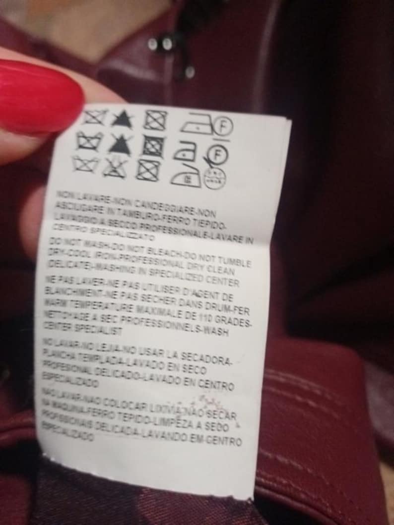 May include: A close-up of a burgundy garment tag with care symbols and text in multiple languages. The tag provides washing, drying, and ironing instructions. The tag is held by a person with red nail polish.