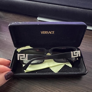 May include: Black sunglasses with a rectangular frame and decorative silver accents on the temples, resting inside a black case. The case is open, revealing a green cleaning cloth and the brand name "VERSACE" in gold lettering.