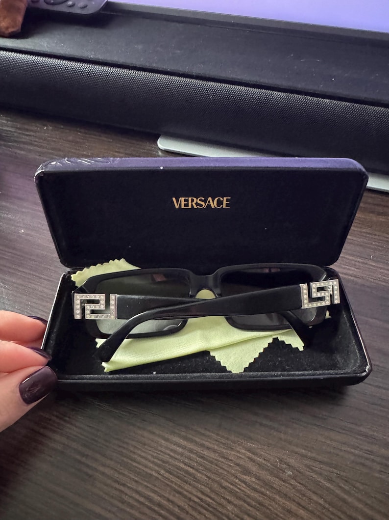 May include: Black sunglasses with a rectangular frame and decorative silver accents on the temples, resting inside a black case. The case is open, revealing a green cleaning cloth and the brand name "VERSACE" in gold lettering.