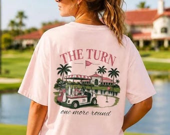 May include: A light pink t-shirt featuring a graphic on the back with the text THE TURN and one more round. The illustration depicts a golf cart parked near a clubhouse, palm trees, and a golf course green with a flag labeled 19.