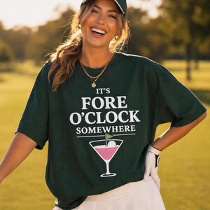 Puede incluir: Una camiseta de manga corta de color verde oscuro con el texto en blanco IT'S FORE O'CLOCK SOMEWHERE sobre un gráfico de una copa de martini rosa que contiene una pelota de golf y una pequeña bandera verde. Cuello redondo y corte holgado.