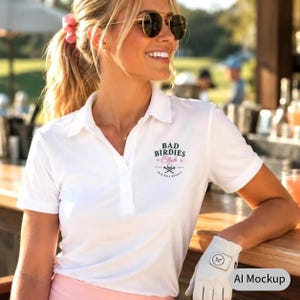 Könnte beinhalten: Ein weißes kurzärmeliges Poloshirt mit Kragen und Knopfleiste. Auf der Brust befindet sich eine Grafik mit dem Text BAD BIRDIES CLUB, einem kleinen Golfball-Symbol und gekreuzten Golfschlägern. Ein weißer Golfhandschuh ist unten rechts zu sehen.