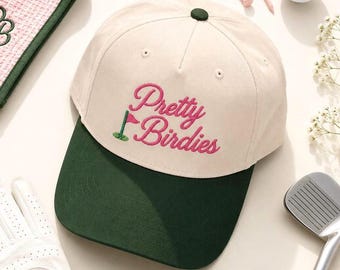 May include: A cream-colored baseball cap with a dark green brim. The front features pink embroidered text that reads Pretty Birdies, accompanied by a small green golf flag icon. The cap has a dark green button at the top center of the crown.