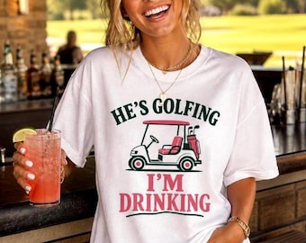 May include: A white short-sleeve t-shirt featuring a graphic of a pink and white golf cart with the text HE'S GOLFING in dark green and I'M DRINKING in pink. The shirt has a crew neckline and a relaxed fit, showcasing a casual style.
