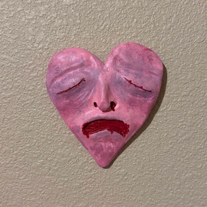 Puede incluir: Una escultura en forma de corazón rosa con una cara, con los ojos cerrados y la boca hacia abajo. La escultura tiene una superficie texturizada y está pintada con tonos de rosa y rojo, transmitiendo una expresión melancólica. La obra de arte está montada en una pared texturizada.