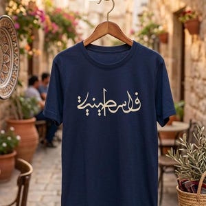 May include: Navy blue t-shirt with cream-colored Arabic script. The shirt is hanging on a wooden hanger against a blurred outdoor background. The text is centered on the front of the shirt.
