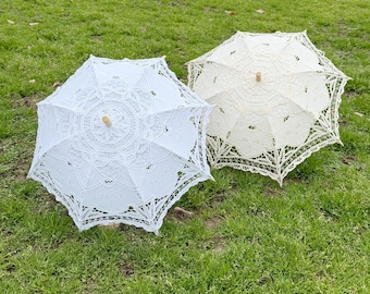 May include: Two parasols featuring intricate white lace patterns with floral motifs and scalloped edges. Each parasol has a central wooden handle and a delicate, openwork design that creates a detailed, circular geometric display.