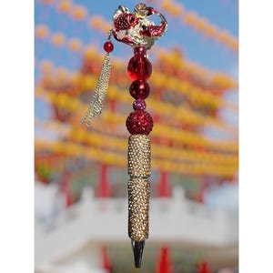May include: A decorative pen with a red and gold design. The pen features a red fish-shaped top, red and gold beads, and a gold-colored, jewel-encrusted barrel. A silver chain with a red bead hangs from the top.