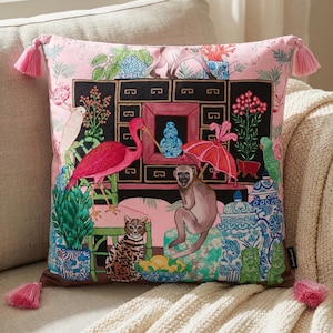 May include: Pink decorative throw pillow with a colorful illustration of a monkey, birds, and other animals in a garden scene. The pillow has pink tassels on each corner and is on a beige couch.