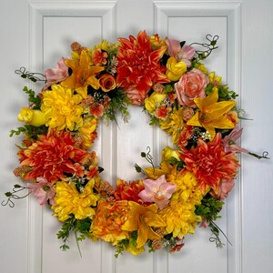 May include: A circular floral wreath featuring a vibrant arrangement of artificial flowers in shades of orange, yellow, and soft pink. The wreath includes large dahlia-like blooms, lilies, and smaller filler flowers interspersed with green foliage.