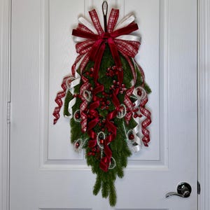 May include: A teardrop-shaped holiday swag featuring green pine branches, clusters of red berries, and a large decorative bow. The bow is crafted from red velvet, white satin, and red-and-white plaid ribbons, with long curling ribbon tails.