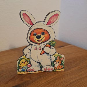Könnte beinhalten: Osterdekoration aus Holz mit einem Bären im Hasenkostüm, der eine Karotte hält. Der Bär ist orange und weiß, mit rosa Akzenten. Ein Küken, ein Hase und Ostereier sind ebenfalls abgebildet.