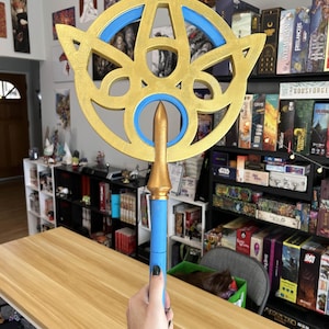 May include: A gold and blue fantasy staff with a star-shaped top. The staff has a blue handle and a gold spearhead. The top has a gold star design with blue accents. The staff is held in front of a wooden table.