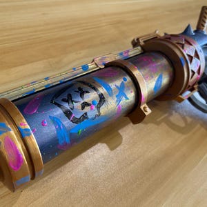 May include: A prop gun with a metallic finish, featuring a skull and crossbones design. The barrel is painted with blue, pink, and gold accents. The handle is black and white.