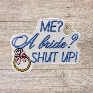 May include: Embroidered patch with the text "ME? A bride? SHUT UP!" in blue script. The patch features a diamond ring with a pink bow. The patch is white with a blue border.