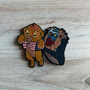 May include: Embroidered patch featuring a cartoon lion cub and a baboon. The lion cub is depicted in shades of brown and tan, with a pink scarf. The baboon is in shades of blue, brown, and red. The patch is likely for clothing or accessories.