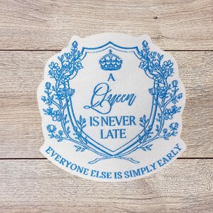 May include: A white patch with a blue floral design and the text "A Queen Is Never Late, Everyone Else Is Simply Early." The design includes a crown and a shield shape.