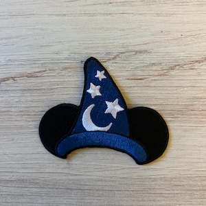 Op de afbeelding: Een blauwe tovenaars hoed patch met witte sterren en een maan ontwerp. De patch heeft zwarte muizenoren aan weerszijden. De patch is waarschijnlijk voor een thema knutselproject of kleding verfraaiing.