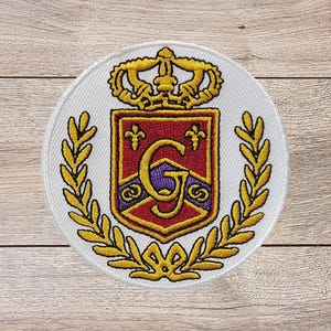 May include: Embroidered patch featuring a heraldic crest design. The crest is red with a gold border and a large letter "G" in the center. A gold crown sits above the crest, and gold laurel branches surround it. The patch is circular with a white background.