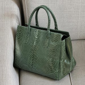 May include: A green handbag with a snakeskin pattern. The bag has two handles and a rectangular shape. The bag is sitting on a gray couch.