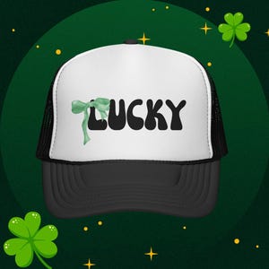 May include: A black and white trucker hat with the word "LUCKY" in black, stylized font. A green bow is on the left side of the word. The hat has a black brim and mesh back. The background is green with shamrocks and stars.