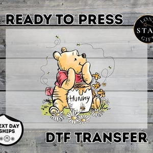 May include: A DTF transfer featuring Winnie the Pooh with a honey pot that says "Hunny". The image includes bees, flowers, and the text "Ready to Press" and "DTF Transfer".