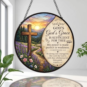 Puede incluir: Un adorno colgante circular estilo vitral que muestra un paisaje con una cruz de madera, un atardecer y flores moradas. El lado derecho muestra el texto: God's God's Grace IS SUFFICIENT FOR THEE, 2 Corinthians 12:9, Joshua 1:9.