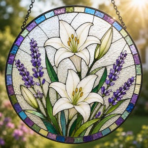 May include: A circular stained glass suncatcher featuring two white lilies with yellow centers and green stems, flanked by purple lavender sprigs. The piece is framed by a border of small, multicolored glass squares and hangs by a metal chain.