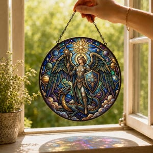 Op de afbeelding: Een ronde glas-in-lood zonnevanger met een gevleugelde figuur in harnas die een speer en schild vasthoudt, staand boven een draak. Het ontwerp bevat een heldere ster, planeten en wolken in tinten blauw, goud en paars, opgehangen aan een ketting.