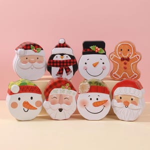 May include: A collection of eight Christmas-themed tins in various shapes. The tins feature designs of Santa Claus, snowmen, a penguin, and a gingerbread man. The color palette includes red, white, and orange, with festive details like holly and plaid patterns.