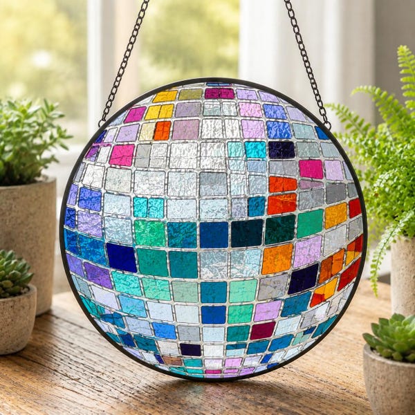 May include: A circular hanging suncatcher featuring a mosaic of small, square, multicolored glass tiles in shades of blue, green, orange, pink, and clear, framed in a thin black metal border and suspended by a black metal chain.