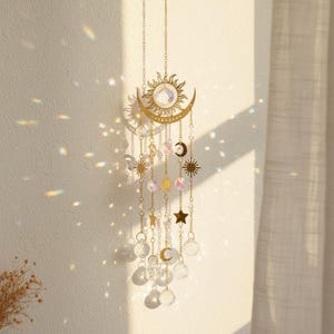 May include: A gold-coloured sun and moon hanging ornament with crystal accents. The celestial-themed decor features a sun with a central crystal, a crescent moon, stars, and dangling crystal beads. The ornament is suspended by a delicate chain.