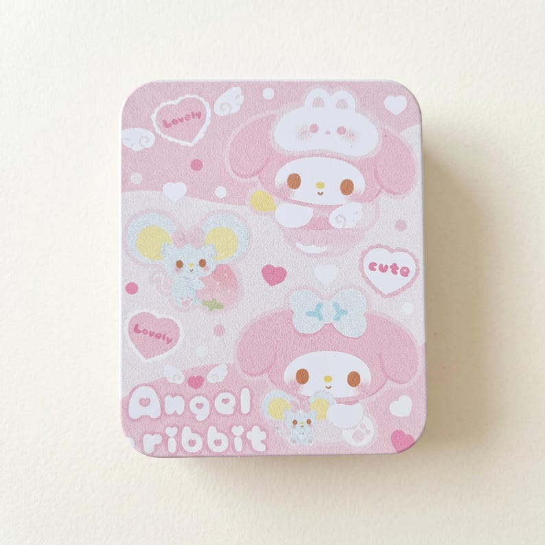 May include: A rectangular pink tin featuring cartoon characters and the text "Angel ribbit." The design includes hearts, clouds, and the word "lovely" in a heart-shaped bubble. The tin has a cute and playful aesthetic.