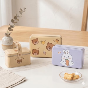 May include: Three rectangular metal boxes with cartoon illustrations. One is light purple with a bunny holding a carrot, another is beige with bear and food graphics, and the third is beige with a handle and bear graphics. A small dish of cookies is in front.