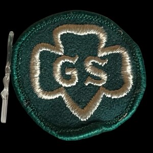 May include: A green embroidered patch featuring a white trefoil design and the letters "GS". The patch has a stitched border and a clear plastic backing.