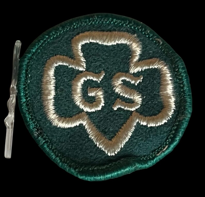 May include: A green embroidered patch featuring a white trefoil design and the letters "GS". The patch has a stitched border and a clear plastic backing.