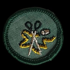 May include: A circular green embroidered patch with a pair of silver scissors, a yellow measuring tape, and a white thread spool. The patch is edged with green. The design is for sewing or tailoring.