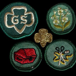 May include: A collection of Girl Scout patches and a gold-colored pin. The patches are round with green backgrounds and embroidered designs, including a trefoil with "GS", flowers, a red box, and scissors. The pin features an eagle.