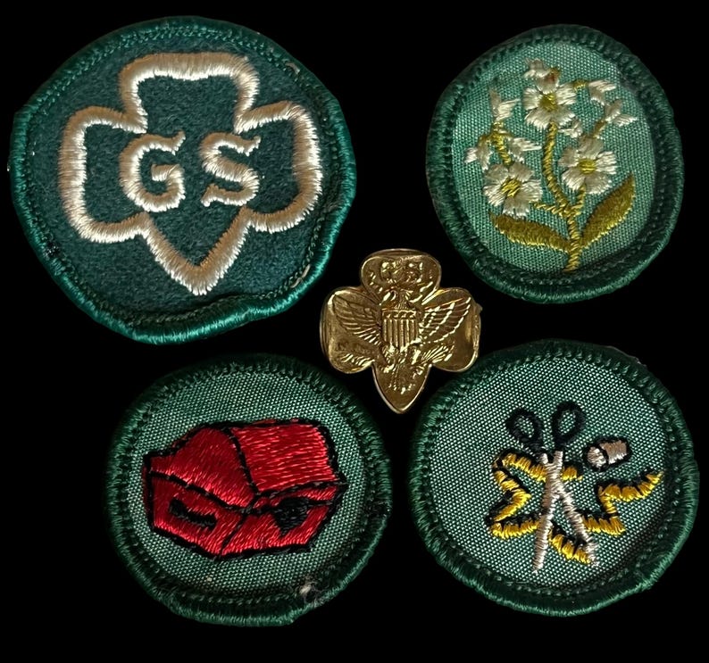 May include: A selection of Girl Guide badges and a gold-coloured pin. The badges are round with green backgrounds and embroidered designs, including a trefoil with "GS", flowers, a red box, and scissors. The pin features an eagle.