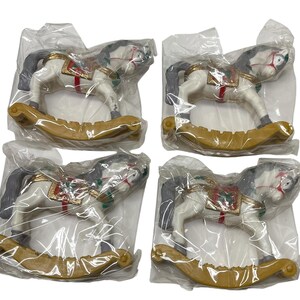 May include: Four white rocking horse figurines with grey manes and tails, each featuring a gold-colored saddle with red and green accents. Each individual horse is enclosed in a clear plastic bag, showing the curved gold-colored rocker base.