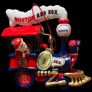 May include: A ceramic Boston Red Sox holiday train ornament featuring Santa Claus in the cabin, a baseball marked 2005, and a gold-toned metal tag reading Boston Red Sox 2005 Holiday Express. The train is painted in red, blue, white, and gold.